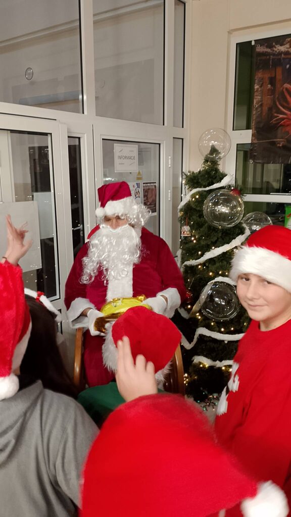 Mikołajki 2025. Na zdjęciu św. Mikołaj siedzi na bujanym fotelu wokół niego są dzieci. W tle choinka. 