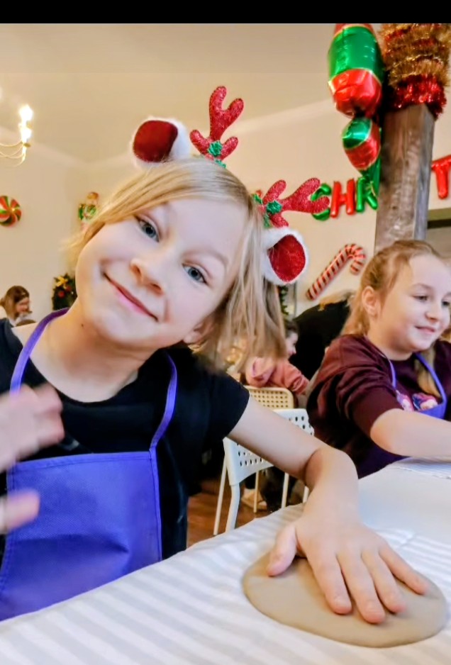 Świąteczne warsztaty ceramiczne w Chichach. Na zdjęciu uczennica uśmiecha się, ma na głowie opaskę z rogami renifera, na stole trzyma rękę na swojej glinie.