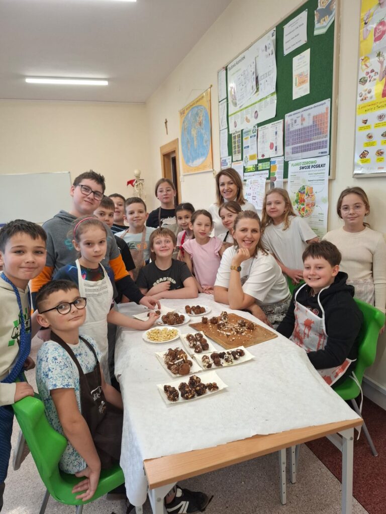 Na zdjęciu uczniowie klasy 4b siedzący przy stole wraz z panią dietetyk i nauczycielką przyrody, prezentujący zrobione podczas zajęć słodkie przekąski.