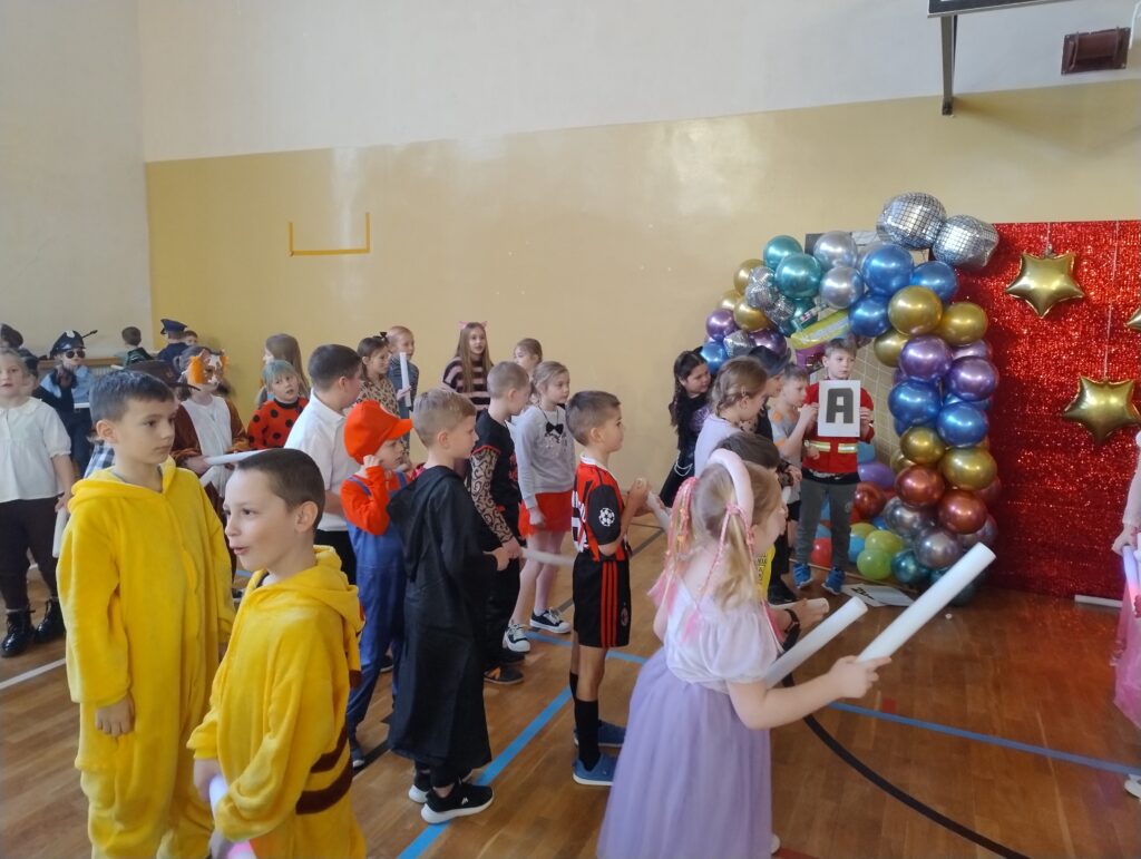 Uczniowie w strojach karnawałowych na sali gimnastycznej podczas jednej z zabaw integracyjnych. W tle kolorowy, balonowy łuk.