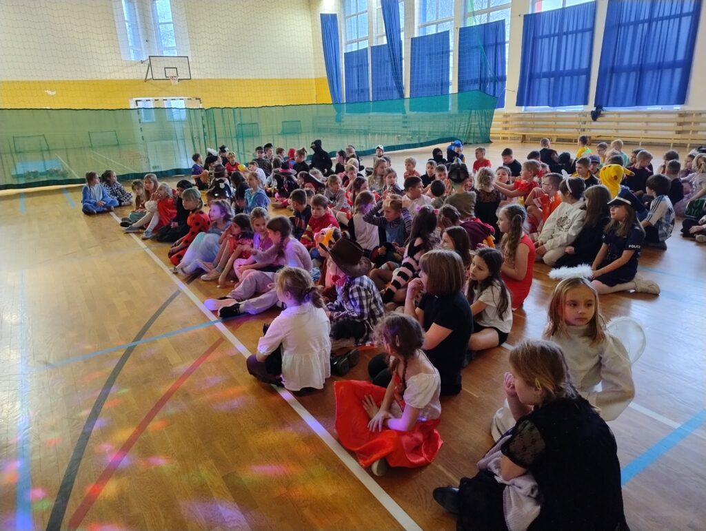 Uczniowie klas 1-3 siedzą na podłodze w sali gimnastycznej i oglądają magiczny pokaz.