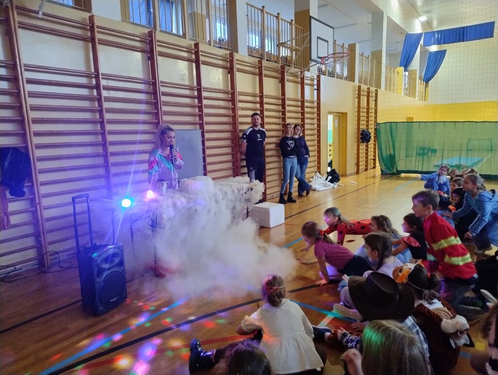 Na zdjęciu widać stanowisko do magicznego pokazu, z którego wydobywa się biały dym. Przed stanowiskiem siedzi grupa dzieci i z zainteresowaniem przygląda się eksperymentowi.