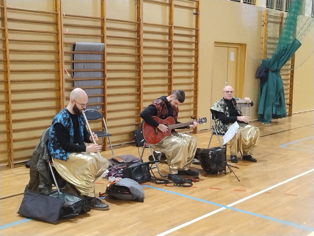 Zdjęcie przedstawia trzech muzyków w strojach stylizowanych na orientalne. Mężczyźni grają na instrumentach – na flecie, na gitarze oraz na dzwonkach wietrznych. 