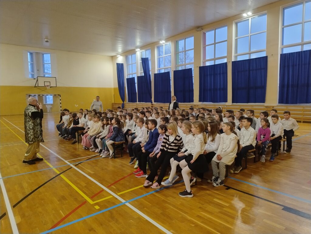 Zdjęcie przedstawia prowadzącego koncertu występującego przez uczniami klas 1-3. Mężczyzna opowiada dzieciom opowieść o Sindbadzie. Uczniowie siedzą na drewnianych ławkach. Są ubrani w galowe stroje – białe koszule oraz ciemne spódniczki i spodnie.