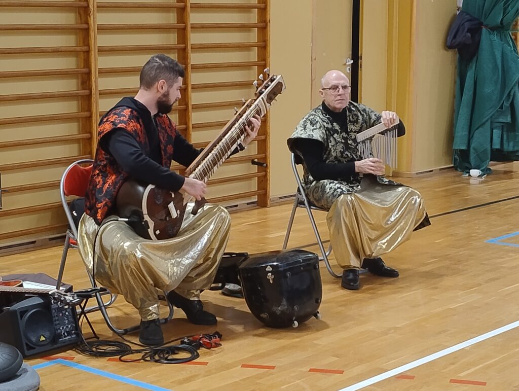 Muzycy siedzą na krzesłach i grają na instrumentach. Młodszy mężczyzna, który siedzi po lewej stronie, gra na sitarze (hinduskiej gitarze), a starszy na dzwonkach wietrznych.