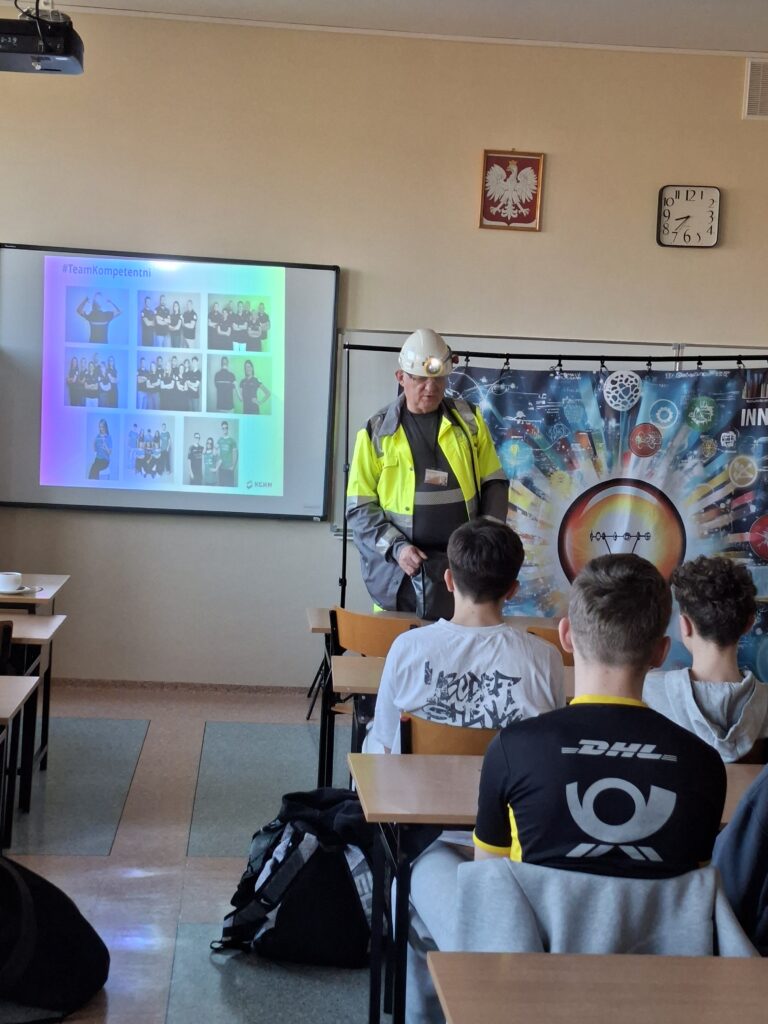 Zdjęcie przedstawia górnika z latarką na głowie,  ubranego w żółtą odblaskową kamizelkę, który opowiada uczniom o swojej pracy. W tle dekoracja i ekran z prezentacją. 
