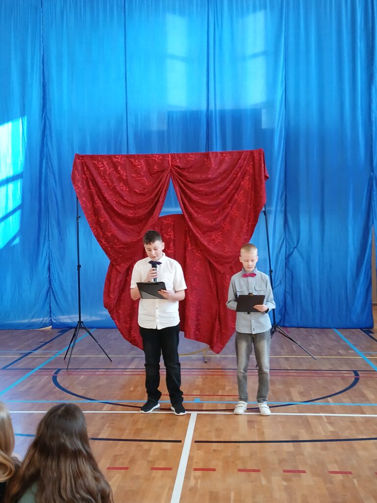 Dzień Kobiet. Na zdjęciu chłopcy z klasy piatej ubrani w a białe koszule prowadzą apel. W tlke czerwony materiał upięty jak kokarda.