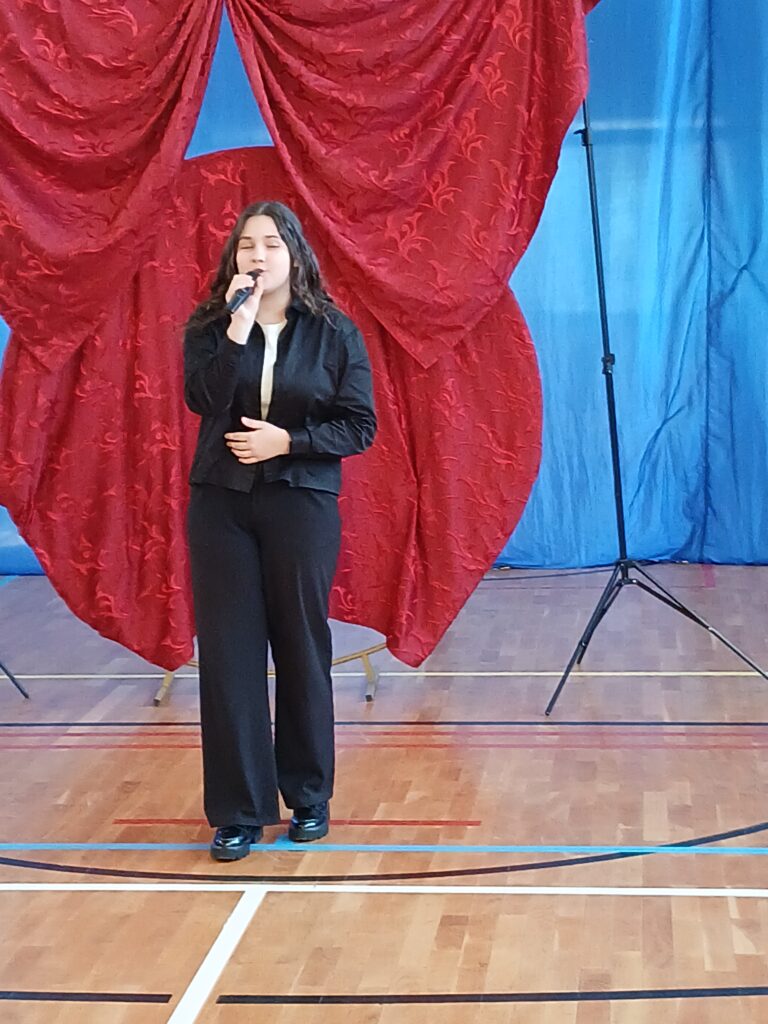 Dzień Kobiet. Na zdjęciu uczennica klasy ósmej ubrana w ciemny garitur śpiea pisenkę. W tle czewrony materiałtworzy dużą kokardę.