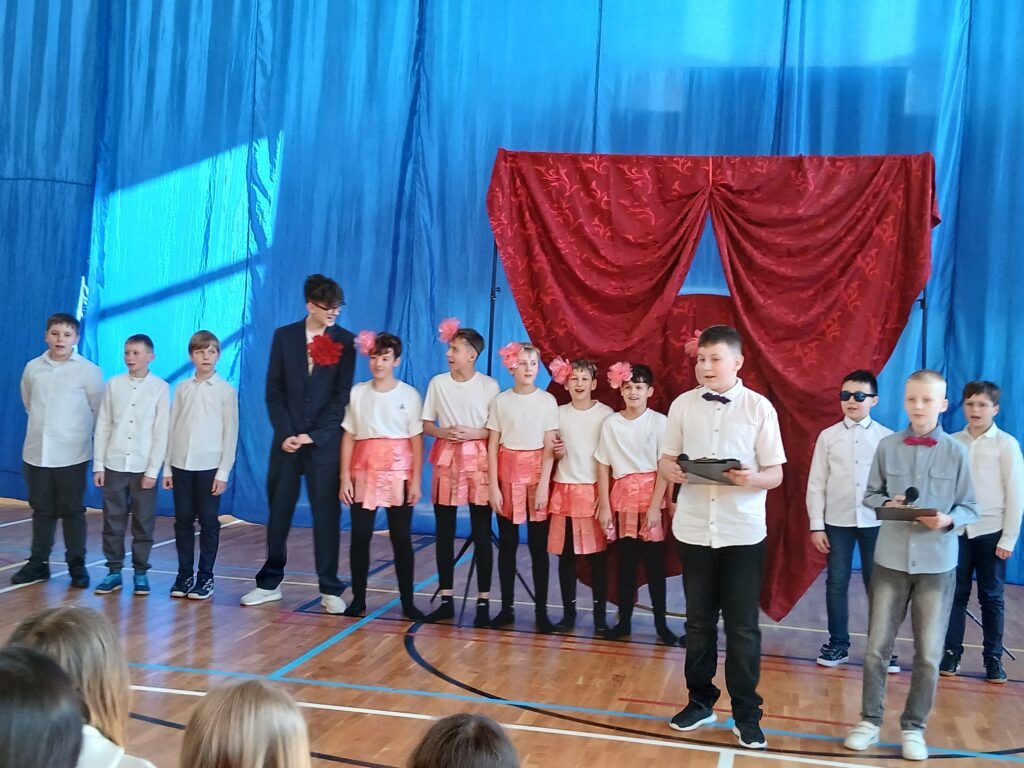 Dzień Kobiet. Na zdjęciu uczniowie z klasy piątej, szóstej, siódmej i ósmej stoją w jednym szeregu i śpiewają STO Lat.