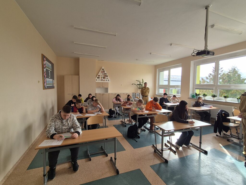 Na zdjęciu widać kilku uczniów siedzących w klasie przy ławkach. Każdy uczeń ma przed sobą arkusz testowy i długopis lub ołówek, skupiony na wypełnianiu zadania. Uczniowie siedzą w rzędach, patrząc w dół na swoje kartki. W tle widać regał z pólkami oraz okno wpuszczające światło dzienne do klasy. Atmosfera jest cicha i skupiona, typowa dla sytuacji egzaminacyjnej.