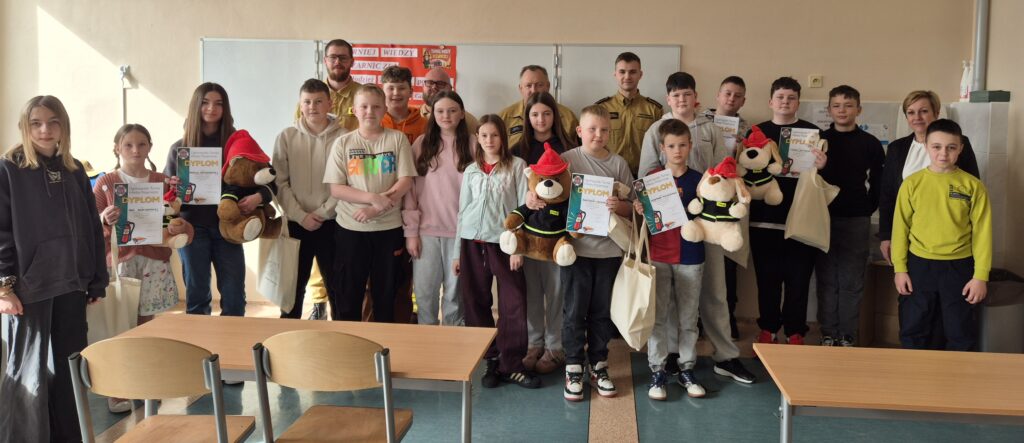 Na zdjęciu widać grupę strażaków stojących razem, Wygrani trzymają w ręku dyplom. Strażacy ubrani są w mundury. Uśmiechają się, a ich postawa jest wyprostowana i pewna siebie. W tle widać salę lub otwartą przestrzeń, ale głównym punktem uwagi są finaliści konkursu i dyplomy, które trzymają. Cała scena to zakończenie konkursu.