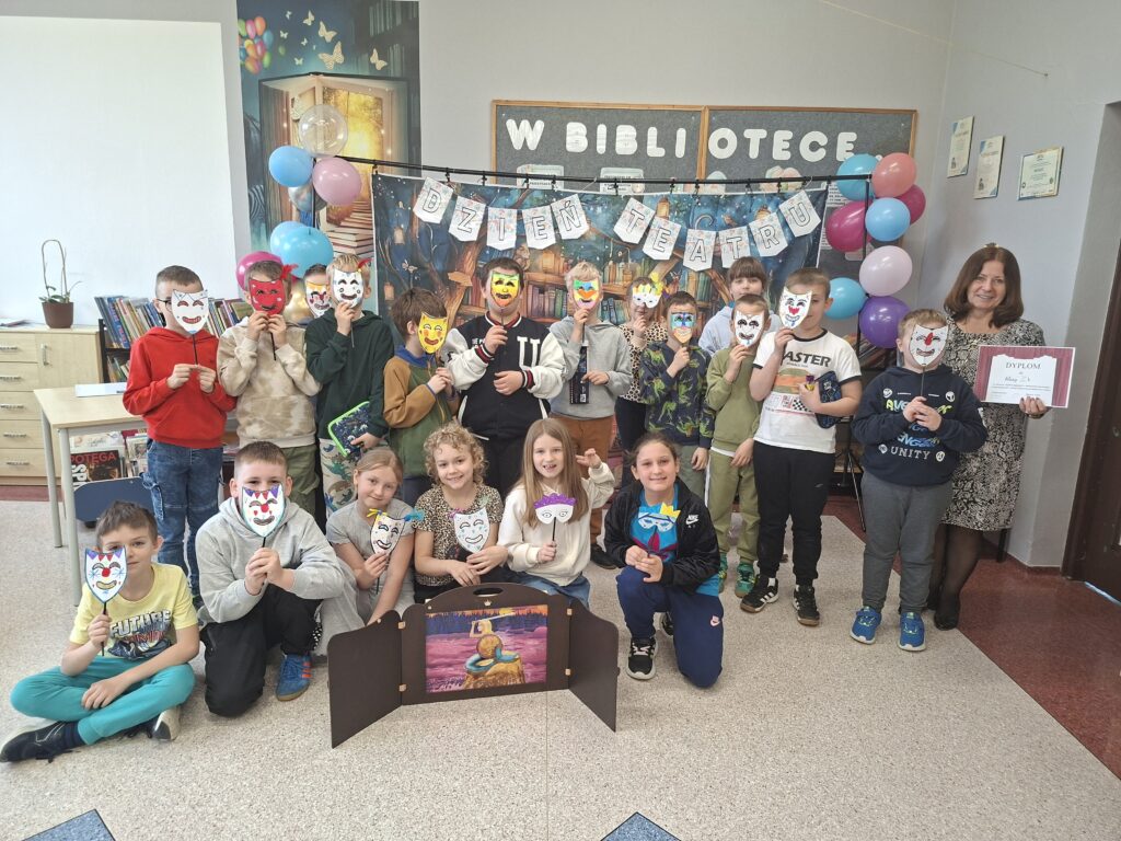 Zdjęcie przedstawia stojących i siedzących uczniów trzymających w ręce udekorowane maski teatralne. Przed nimi stoi teatrzyk kamishibai, czyli drewniana, otwierana skrzynka z kolorowymi kartami opowieści oraz kolorowe poduszki do siedzenia. W tle kolorowy napis "Dzień teatru" udekorowany kolorowymi balonami.