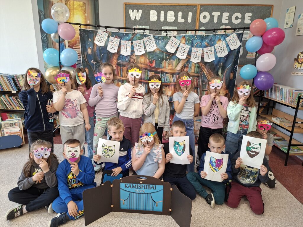 Zdjęcie przedstawia stojących uczniów trzymających w ręce udekorowane maski teatralne. Przed nimi stoi teatrzyk kamishibai, czyli drewniana, otwierana skrzynka z kolorowymi kartami opowieści. W tle kolorowy napis "Dzień teatru" udekorowany kolorowymi balonami.