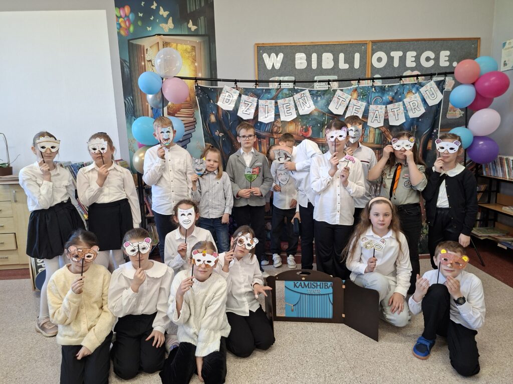 Zdjęcie przedstawia stojących uczniów trzymających w ręce udekorowane maski teatralne. Przed nimi stoi teatrzyk kamishibai, czyli drewniana, otwierana skrzynka z kolorowymi kartami opowieści. W tle kolorowy napis "Dzień teatru" udekorowany kolorowymi balonami.