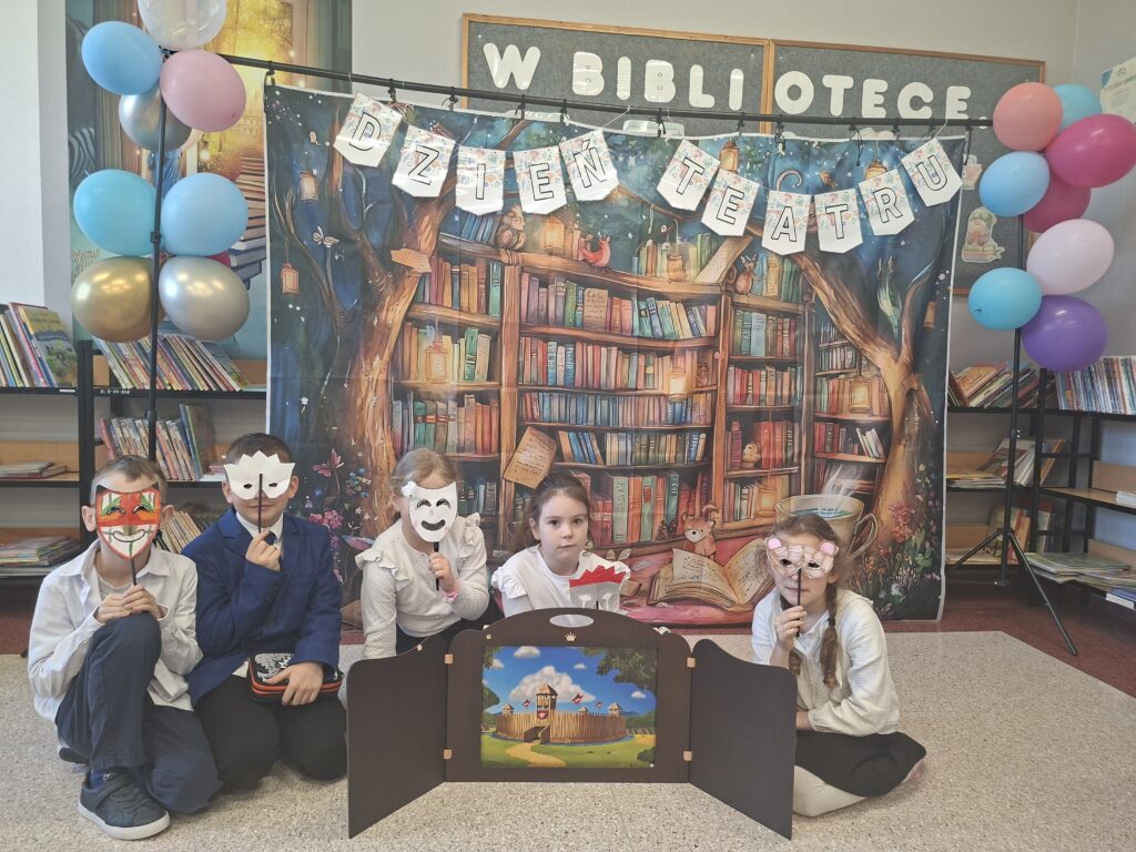 Zdjęcie przedstawia siedzących uczniów trzymających w ręce udekorowane maski teatralne. Przed nimi stoi teatrzyk kamishibai, czyli drewniana, otwierana skrzynka z kolorowymi kartami opowieści. W tle kolorowy napis "Dzień teatru" udekorowany kolorowymi balonami.