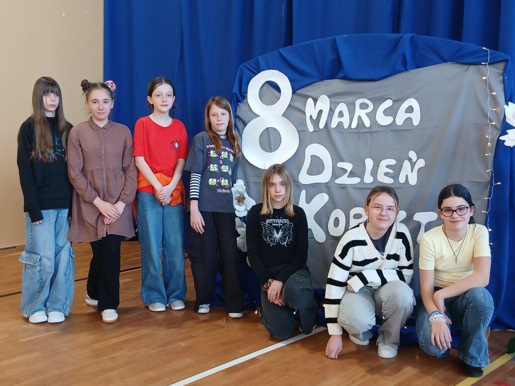 Uczennice klas piątych i szóstych pozują do zdjęcia przy tablicy, na której znajduje się dekoracja: papierowe białe kwiaty i napis 8 marca Dzień Kobiet.