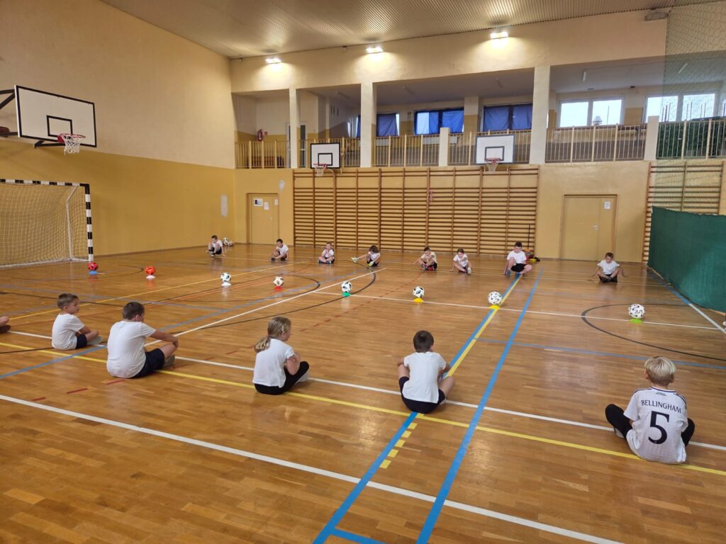 Program "Aktywna Szkoła do Kwadratu". Na zdjęciu uczniwoie siedzą na przecie siebie na sali gimnastycznej. Posródku nich jest piłka. Po prawej stronie stoi bramka do pilki nożnej.