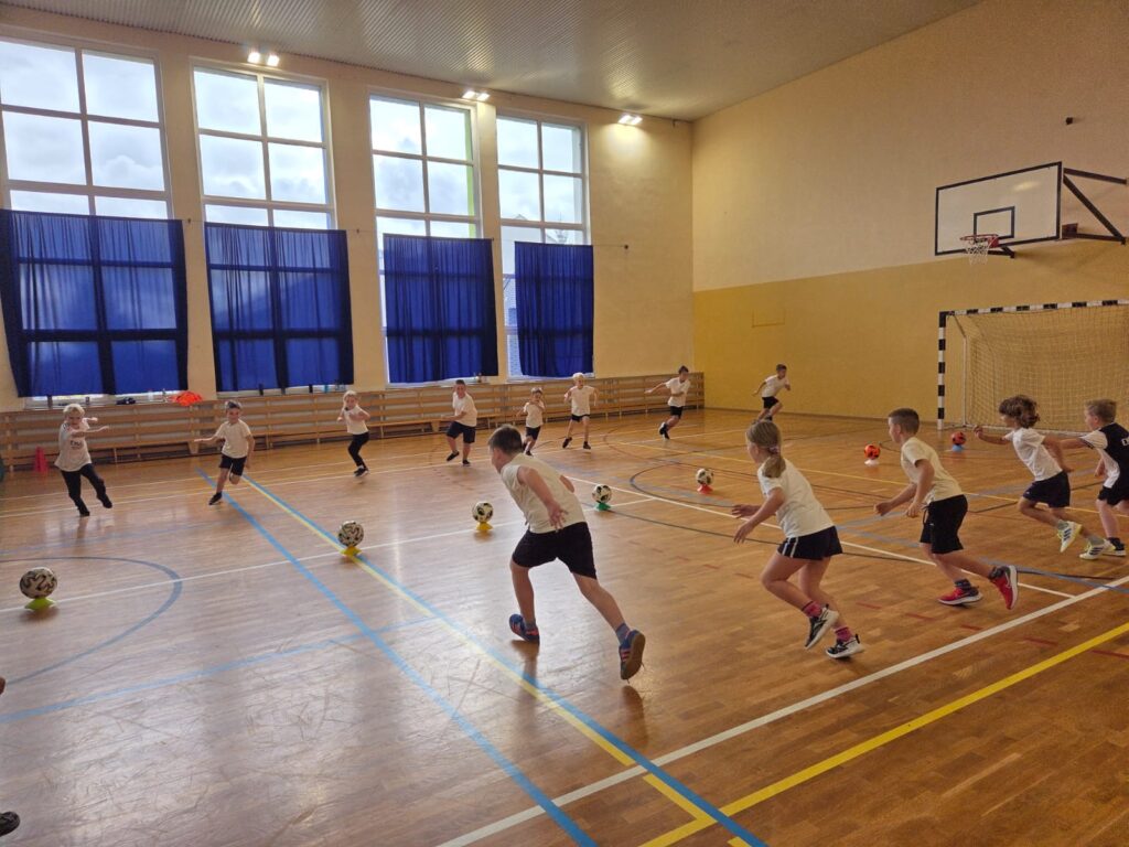 Program "Aktywna Szkoła do Kwadratu". Na zdjęciu uczniwoie wykonują ćwiczenie z piłką, biegną i omijają piłkę.