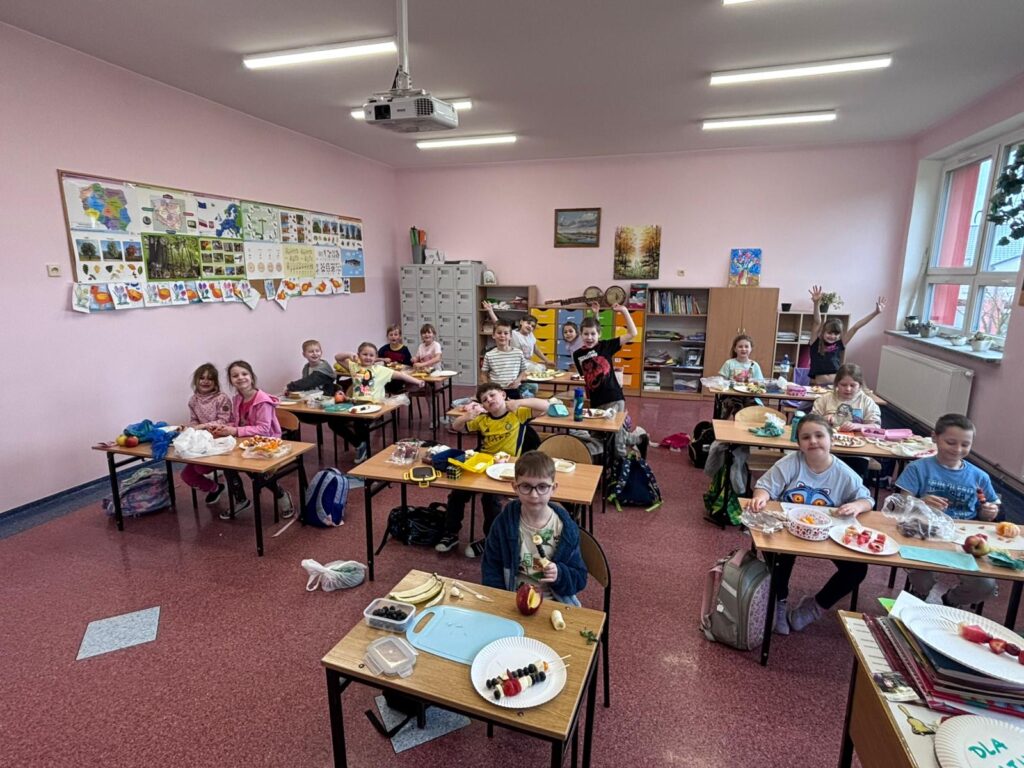 Uzniowie klasy 1a znajdują się w swojej sali lekcyjnej. Siedzą w ławkach lub stoją przy nich i przygotowują kanapki oraz owocowe szaszłyki.
