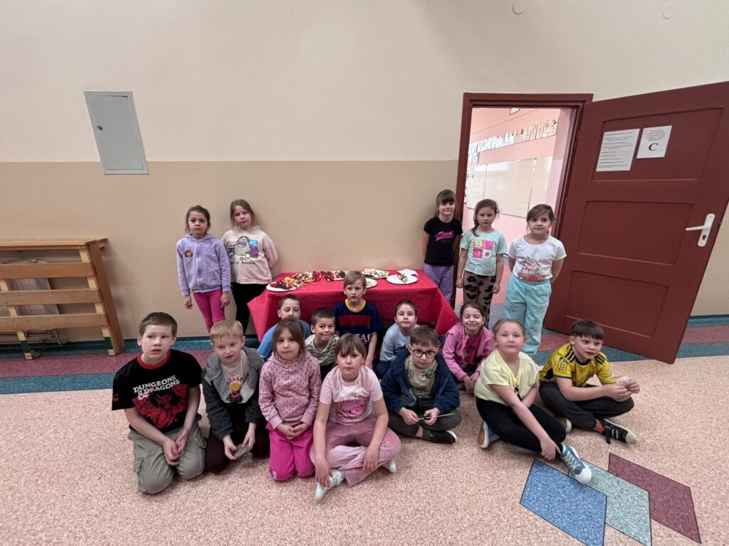 Uczniowie klasy 1a pozują do zdjęcia z przygotowanymi przed siebie zdrowymi przekąskami. Kanapki i szaszłyki są podane na stoliku przed wejściem do klasy.
