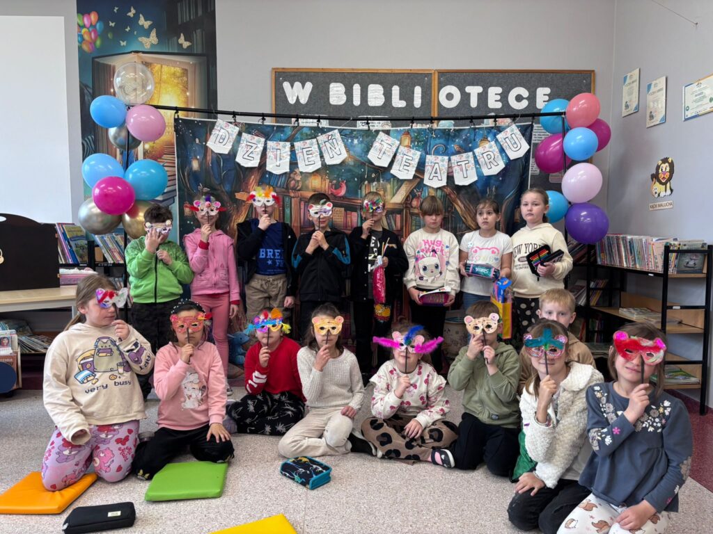 Zdjęcie przedstawia stojących uczniów trzymających w ręce udekorowane maski teatralne. Przed nimi stoi teatrzyk kamishibai, czyli drewniana, otwierana skrzynka z kolorowymi kartami opowieści. W tle kolorowy napis "Dzień teatru" udekorowany kolorowymi balonami.