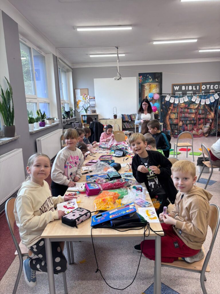 Zdjęcie przedstawia siedzących uczniów trzymających przt stołach wykinujących kolorowe maski. Na stołach leżą piórniki z przyborami szkolnymi.