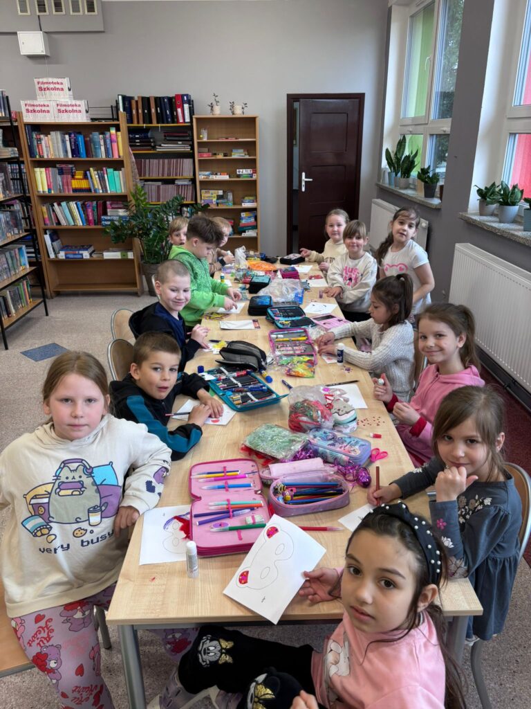 Zdjęcie przedstawia siedzących uczniów trzymających przt stołach wykinujących kolorowe maski. Na stołach leżą piórniki z przyborami szkolnymi.