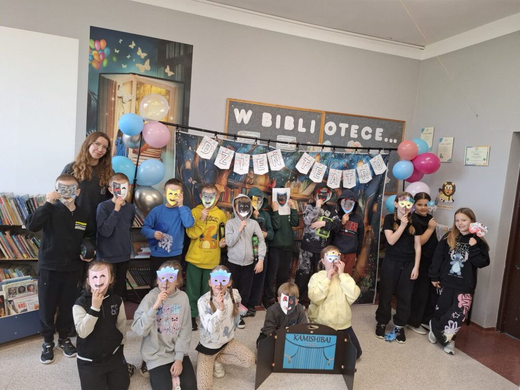Zdjęcie przedstawia stojących uczniów trzymających w ręce udekorowane maski teatralne. Przed nimi stoi teatrzyk kamishibai, czyli drewniana, otwierana skrzynka z kolorowymi kartami opowieści. W tle kolorowy napis "Dzień teatru" udekorowany kolorowymi balonami.