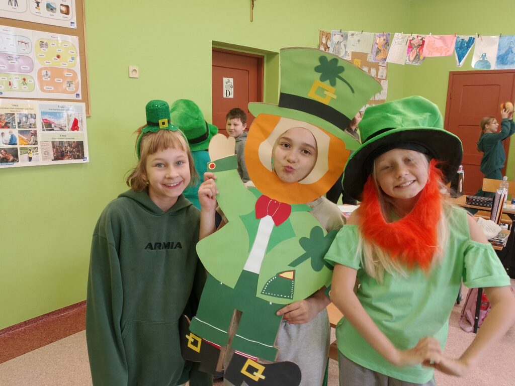 Obchody Dnia św. Patryka- patrona Irlandii. Na zdjęciu trzy uczennice pozują do zdjęcia. Jedna ma na głowie zielony kapelusz, druga trzyma wyciętego z papieru skrzata Leprechauna, a trzecia ma na głowie zieloną opaskę.