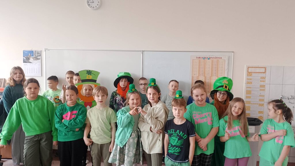 Obchody Dnia św. Patryka- patrona Irlandii. Na zdjeciu uczniowie klasy 2a ubrani w elementy stroju w kolorze zielonym pozują do zdjęcia. Dwoje uczniów ma na sobie zielony kapelusz, trzy uczennice mają na głowie zielone opaski. Wszyscy uczniowie trzymają w ręce małego wykonanego z papieru skrzata Leprechaun.