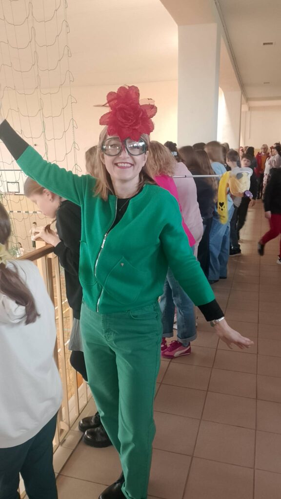 Na zdjęciu nauczycielka bilogii ubrana całą na zielono, na głowie ma czerwony kwiatek a na twarzy duże okulary z zielonymi oprawkami. 