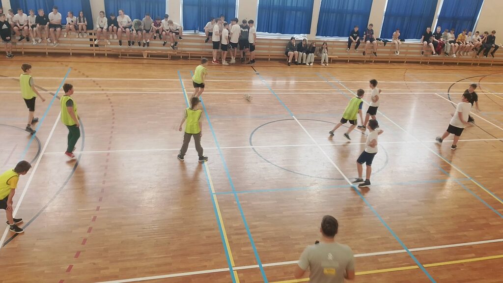 Na zdjęciu uczniowie klas czwartcyh podzczas rozgrywek w piłkę nożną. Niektórzy uczniwoie mają na sobie żólte kamizelki. W tle widać innych uczniów z klas 4-6. 