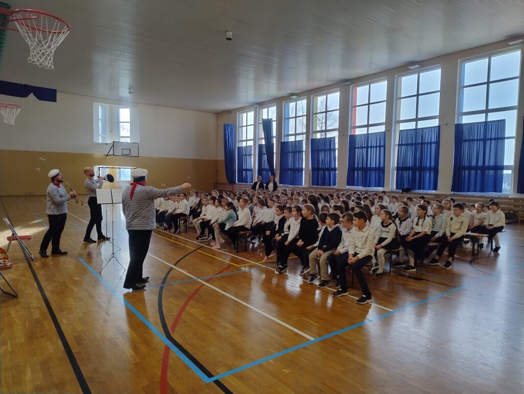 Zdjęcie przedstawia trzech muzyków w kostiumach marynarzy. Mężczyźni występują przed uczniami klas 1-3, siedzącymi na widowni. Uczniowie są ubrani w stroje apelowe.