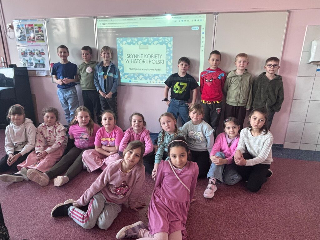 Zdjęcie przedstawia uczniów klasy 1a pozujących w grupie, na tle tablicy z wyświetloną prezentacją na temat znanych Polek. W pierwszym rzędzie siedzą dziewczynki ubrane na różowo, za nimi stoją chłopcy.