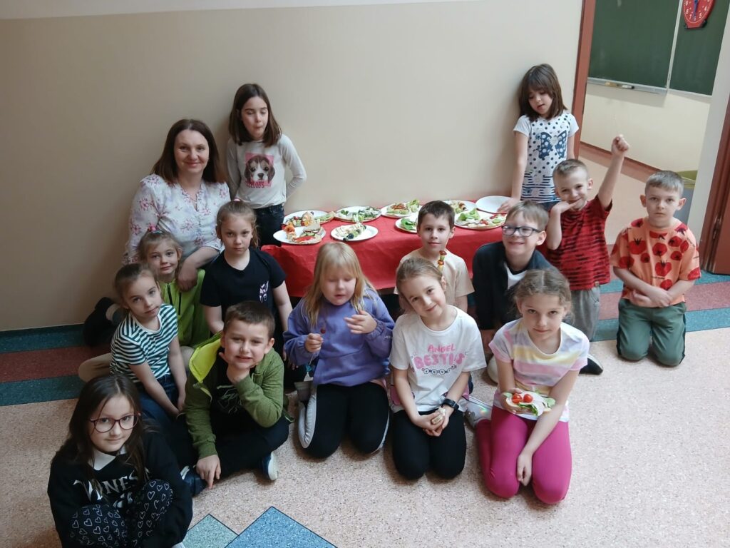 Uczniowie klasy 1b ze swoją wychowawczynią pozują do zdjęcia z przygotowanymi przed siebie zdrowymi przekąskami. Kanapki i szaszłyki są podane na stoliku przed wejściem do klasy.
