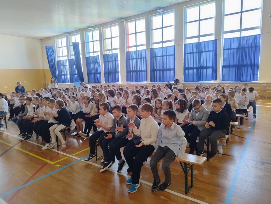 Uczniowie ubrani w stroje apelowe siedzą na drewnianych ławkach i klaszczą i rytm granej muzyki, aktywnie uczestnicząc w koncercie.
