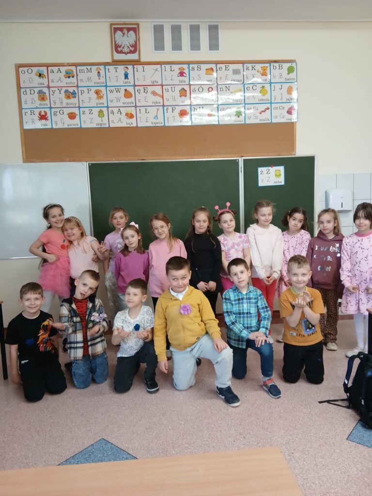 Zdjęcie przedstawia uczniów klasy 1b pozujących w grupie w swojej klasie. Chłopcy w pierwszym rzędzie, a za nimi dziewczynki ubrane na różowo.
