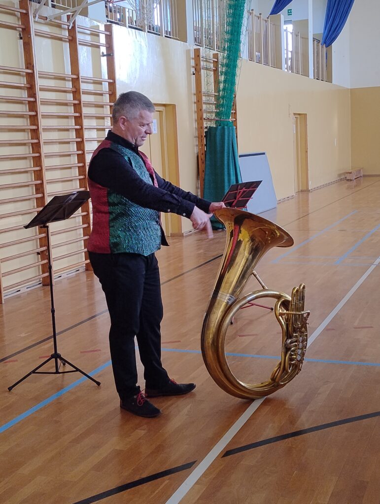 Zdjęcia przedstawia muzyka – mężczyznę w ciemnych spodniach i czerwono-zielonej kamizelce, który prezentuje dzieciom helikon – największy instrument z rodziny instrumentów dętych.
