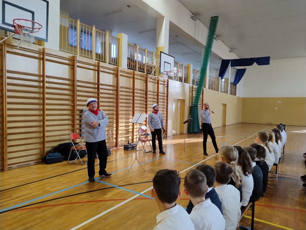 Zdjęcie przedstawia muzyków – tym razem od frontu. Są ubrani w czarne spodnie, koszulki w biało-granatowe paski oraz czerwone chustki i czapki marynarskie. Mężczyzna po prawej stronie zdjęcia trzyma w ręku mikrofon i opowiada dzieciom historię pana Maluśkiewicza.