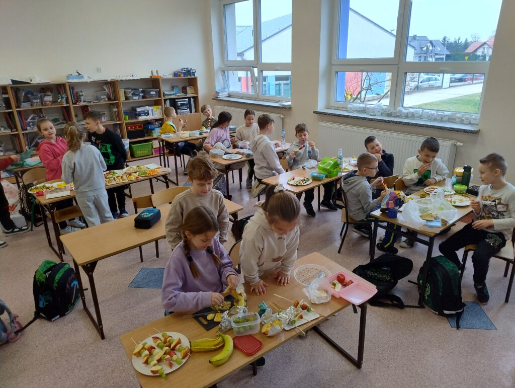 Uczniowie klasy 2a znajdują się w swojej sali lekcyjnej. Podzieleni na małe grupki siedzą w ławkach lub stoją przy nich i przygotowują kanapki oraz owocowe szaszłyki.
