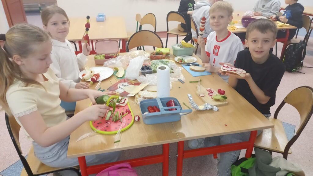  Zdjęcie przedstawia kilkoro uczniów z klasy 2b. Dzieci siedzą przy dwóch połączonych ławkach i przygotowują zdrowe przekąski.
