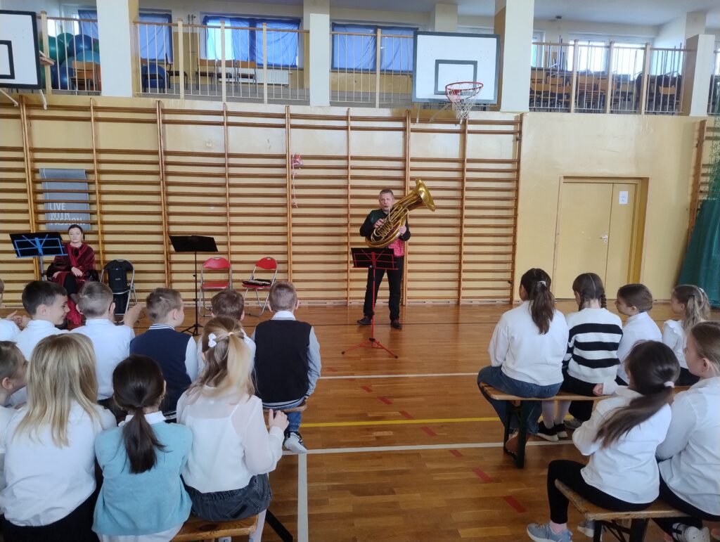 Zdjęcia przedstawia muzyka – mężczyznę w ciemnych spodniach i czerwono-zielonej kamizelce, który prezentuje dzieciom helikon – największy instrument z rodziny instrumentów dętych.
