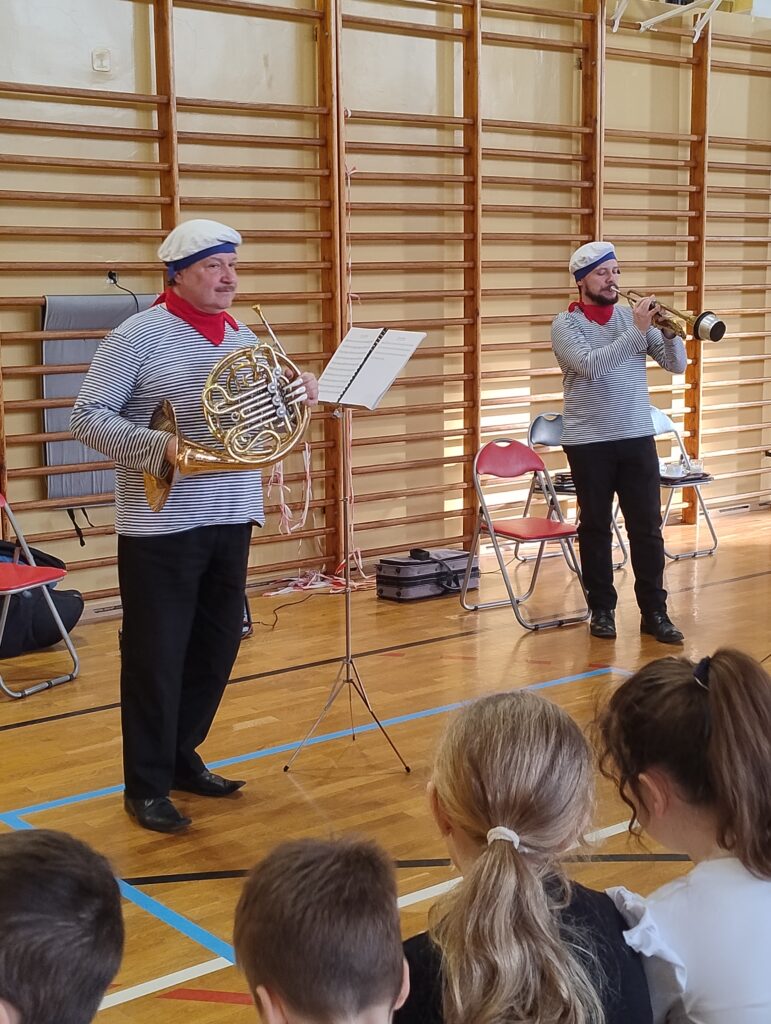 Zdjęcie przedstawia dwóch muzyków w kostiumach marynarzy. Mężczyzna po lewej stronie trzyma w rękach waltornię (róg francuski), a mężczyzna po prawej stronie gra na trąbce z zamontowanym tłumikiem.