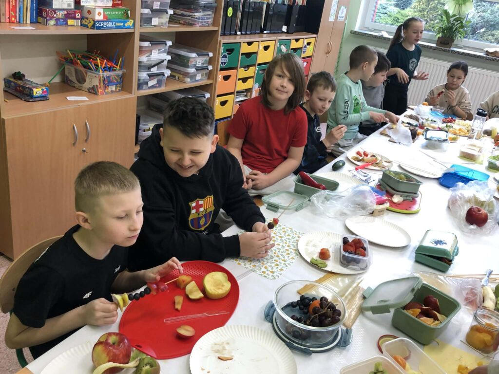 Zdjęcie przedstawia uczniów klasy 3a siedzących przy złączonych stolikach i przygotowujących zdrowe przekąski.
