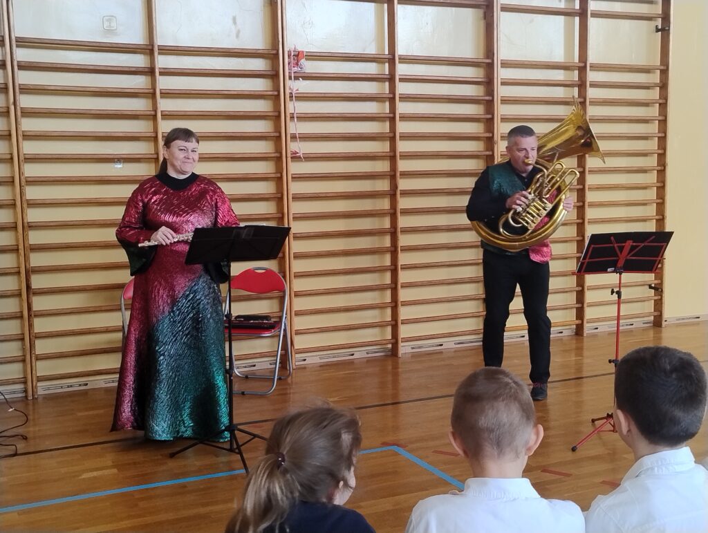 Zdjęcie przedstawia muzyków. Po lewej stronie stoi kobieta w długiej sukni. Trzyma w rękach flet piccolo i się uśmiecha. Po prawej stronie stoi mężczyzna i gra na helikonie.

