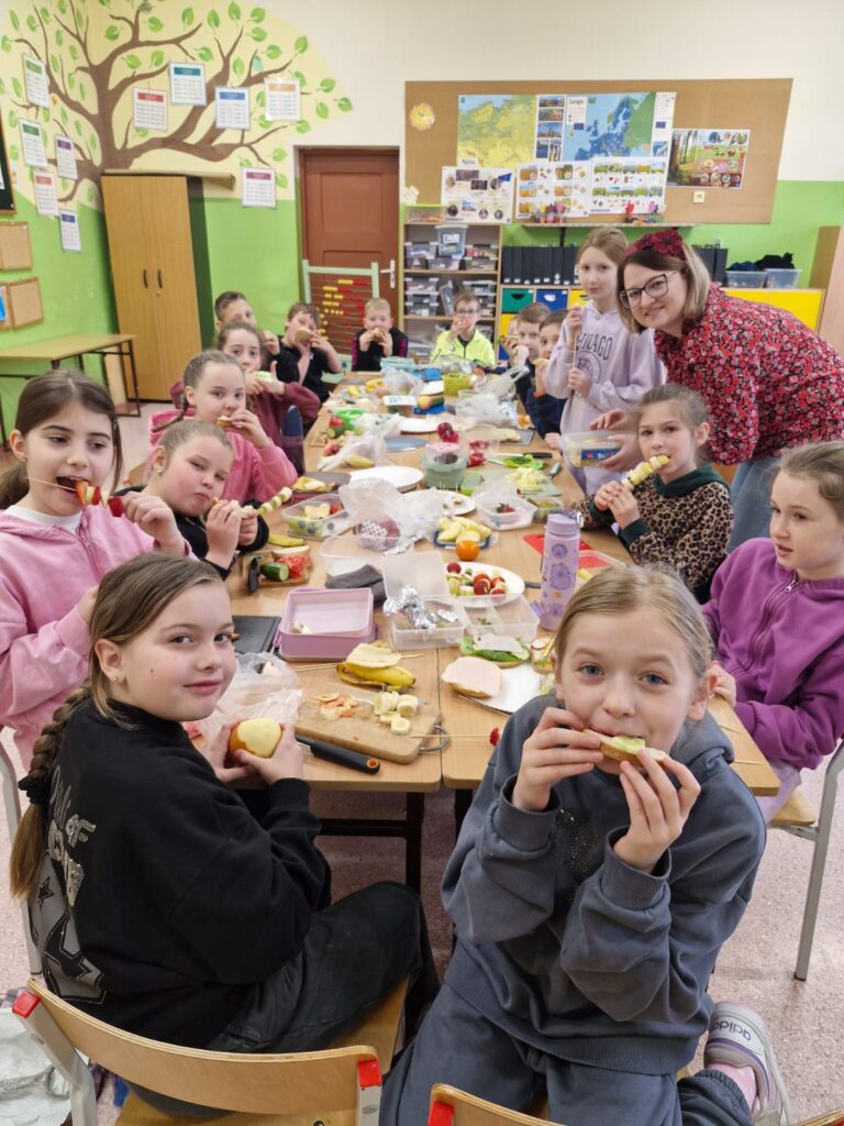 Na zdjęciu znajduje się cały oddział klasowy 3b. Dzieci siedzą przy wspólnym stole i zajadają się przygotowanymi przez siebie kanapkami oraz owocowymi szaszłykami.
