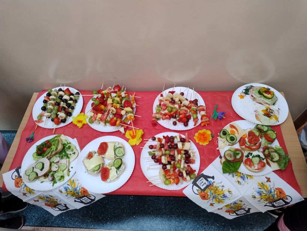 a zdjęciu widoczna jest jedna ze stacji ze zdrowymi przekąskami przygotowanymi przez uczniów. Na ławce przykrytej czerwonym obrusem i wiosennymi serwetkami znajdują się talerzyki wypełnione kolorowymi kanapkami i szaszłykami.
