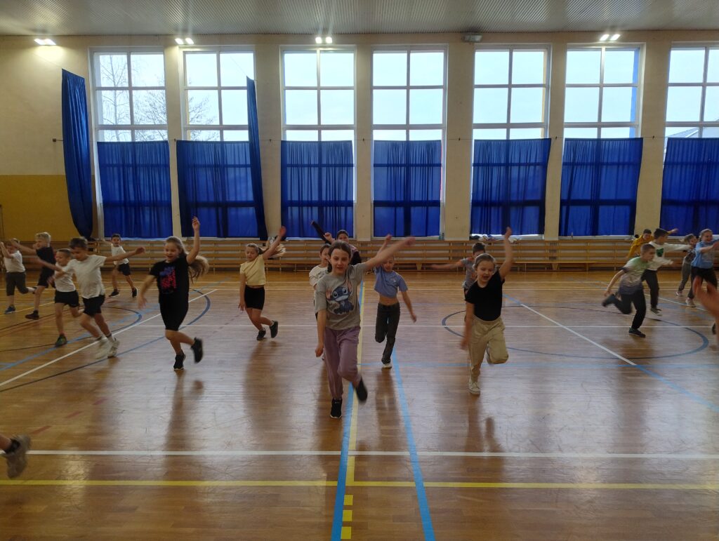 Zdjęcie przedstawia uczniów klas drugich i trzecich w trakcie rozgrzewki przed zajęciami sportowymi. Uczniowie biegną w kierunku obiektywu, naprzemiennie wymachując ramiona.
