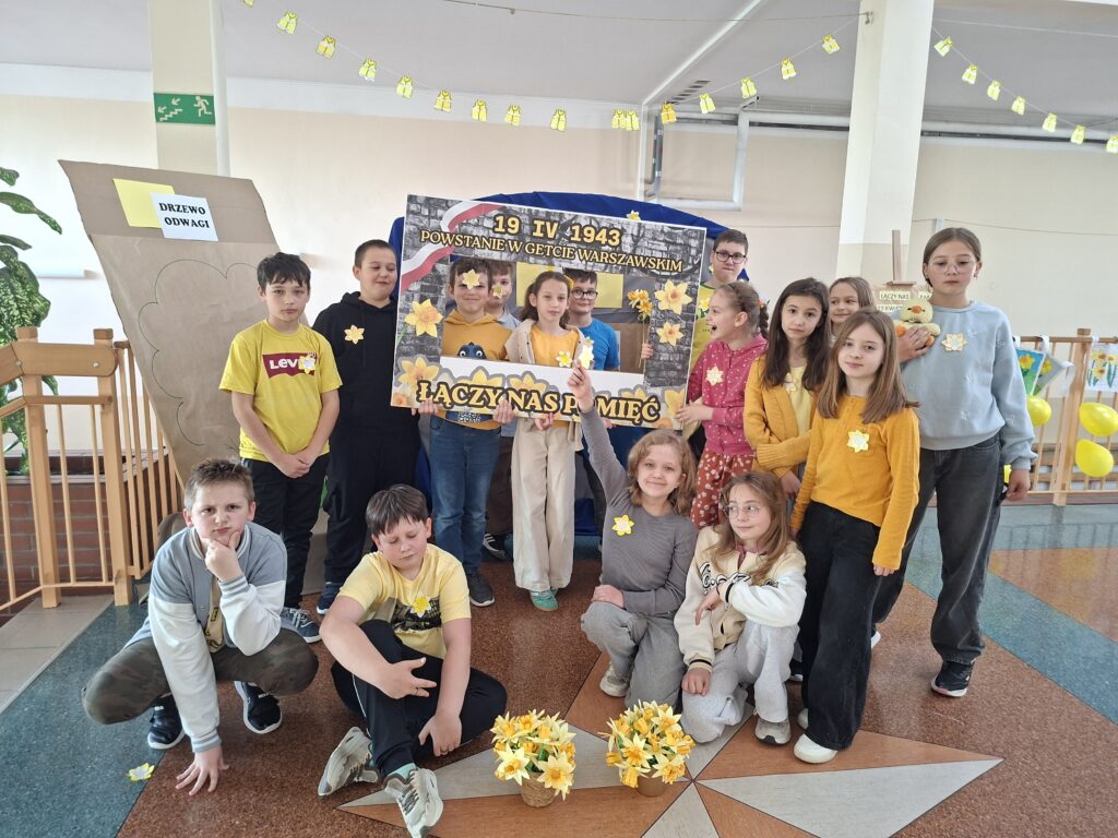 Zdjęcie przedstawia uczniów ubranych w kolorze żółtym z fotobudką - żółty napisz "Łączy nas pamięć". Przed uczniami stoją wazony z żółtymi żonkilami. W tle narysowane "drzewo odwagi".