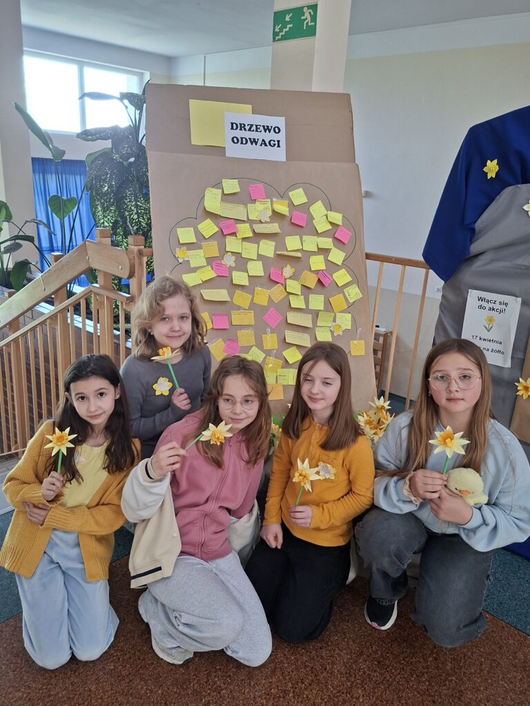 Dziewczynki na szkolnym korytarzu trzymają w ręce żółte żonkile. Za nimi "Drzewko odwagi" z naklejonymi żółtymi i różowymi karteczkami z wyjaśnieniem, czym jest dla mnie odwaga.